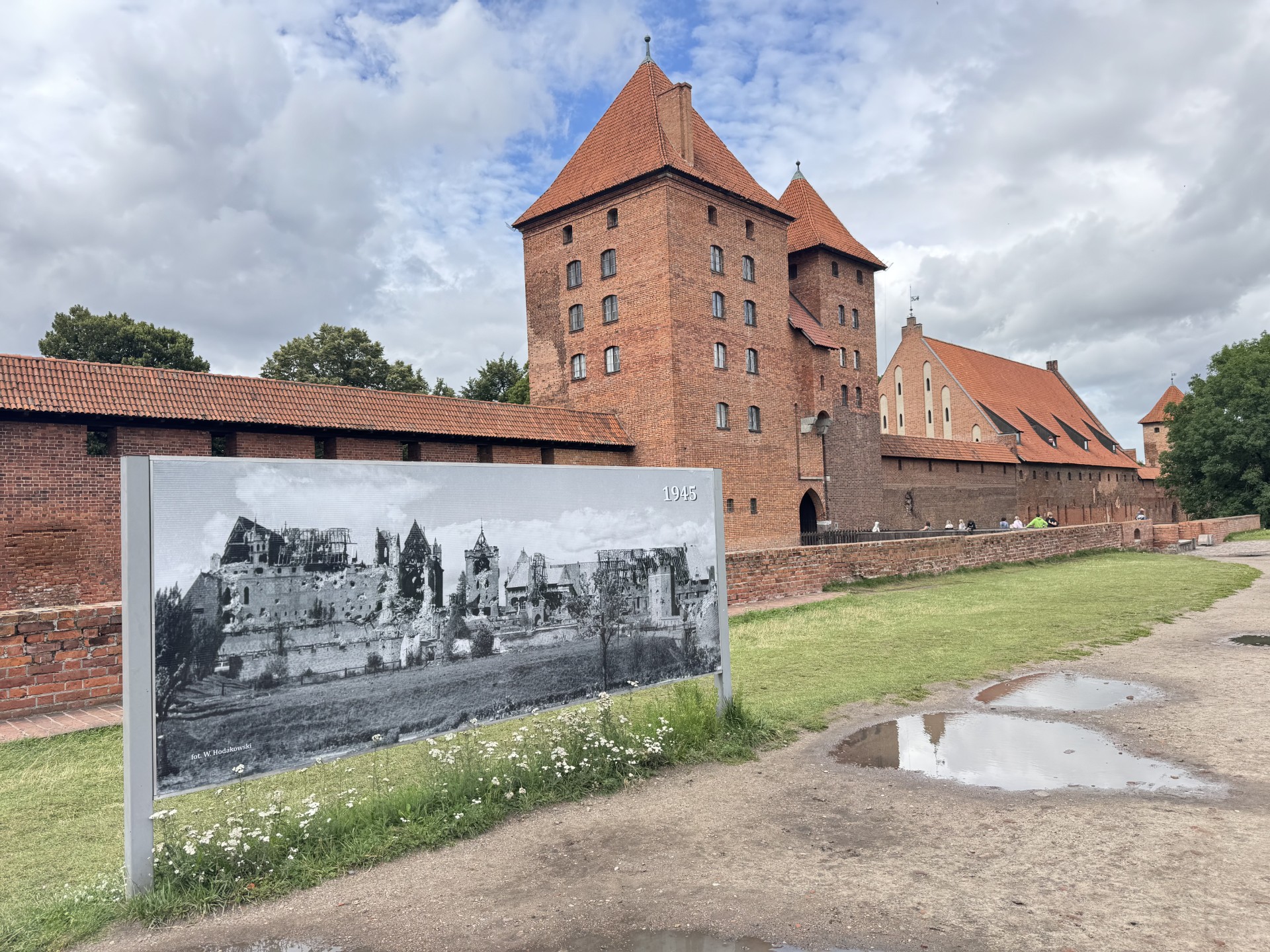 Malbork Castle in Poland, a red-brick medieval Teutonic fortress, with a black-and-white 1945 photo panel in the foreground showing the castle in wartime ruins.